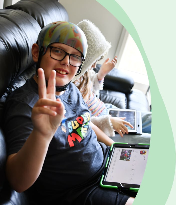 A young boy is showing a peace sign whilst playing his ipad