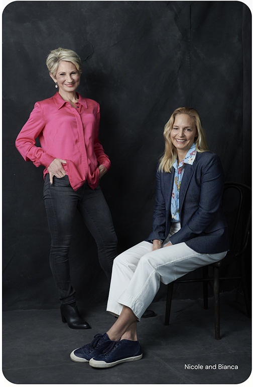 Nicole standing with her hands in her pockets and Bianca sitting on a chair with her legs extended and crossed. Both look relaxed and are smiling.