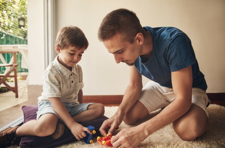 Dad sits on floor with young son and plays with toy trains