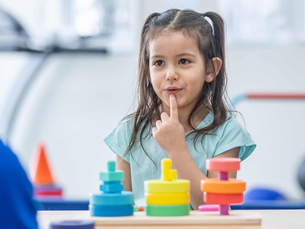 Young girl with pigtails touching her lips thoughtfully while sitting at a table with colorful stacking toy towers, appearing to concentrate on speech sounds.
