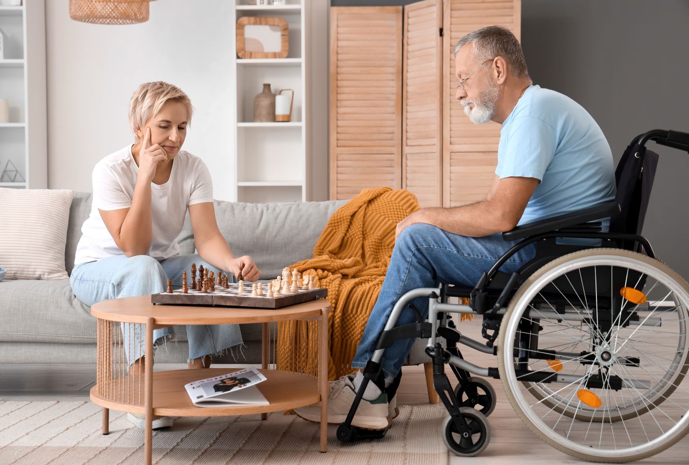 Man seated in wheelchair playing chess with woman seated on couch