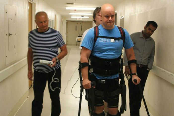 A paralyzed patient walks using an exoskeleton and assistance from transcutaneous spinal cord stimulation. Three men stand behind and assist him 