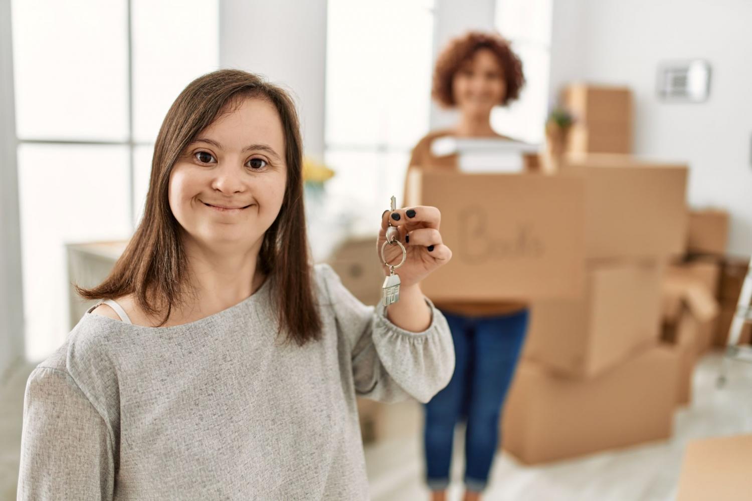 Woman holding up key with carer in background holding and surrounded by moving boxes