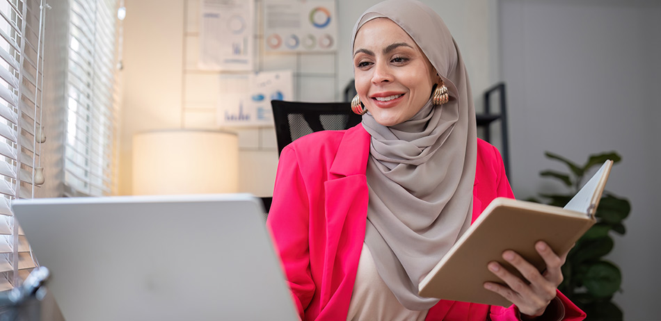 Young woman smiling whilst looking at her notebook and adding info in her laptop