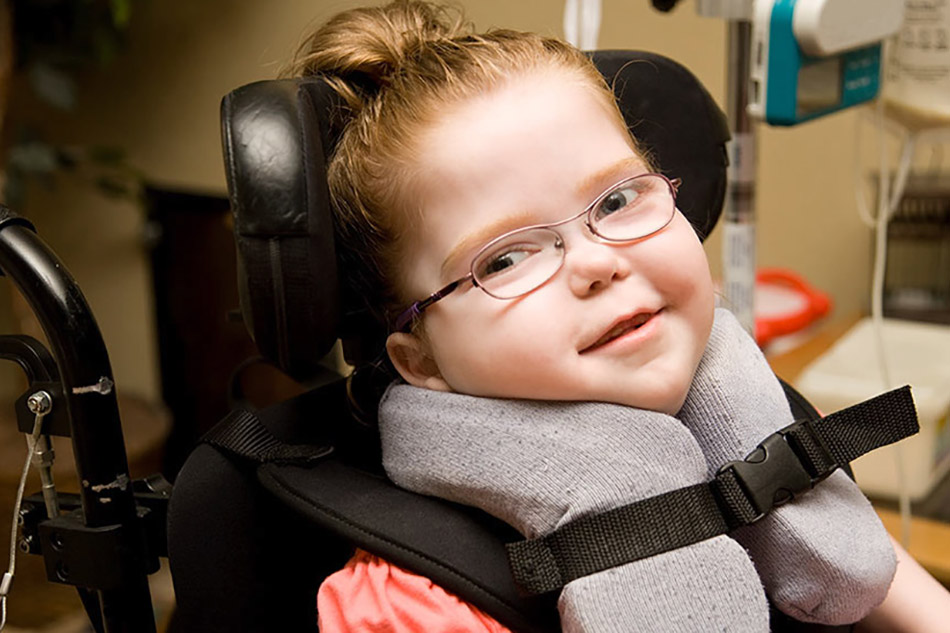 A gorgeous little girl sitting in a supported wheelchair. She has a pony tail and super cute glasses.