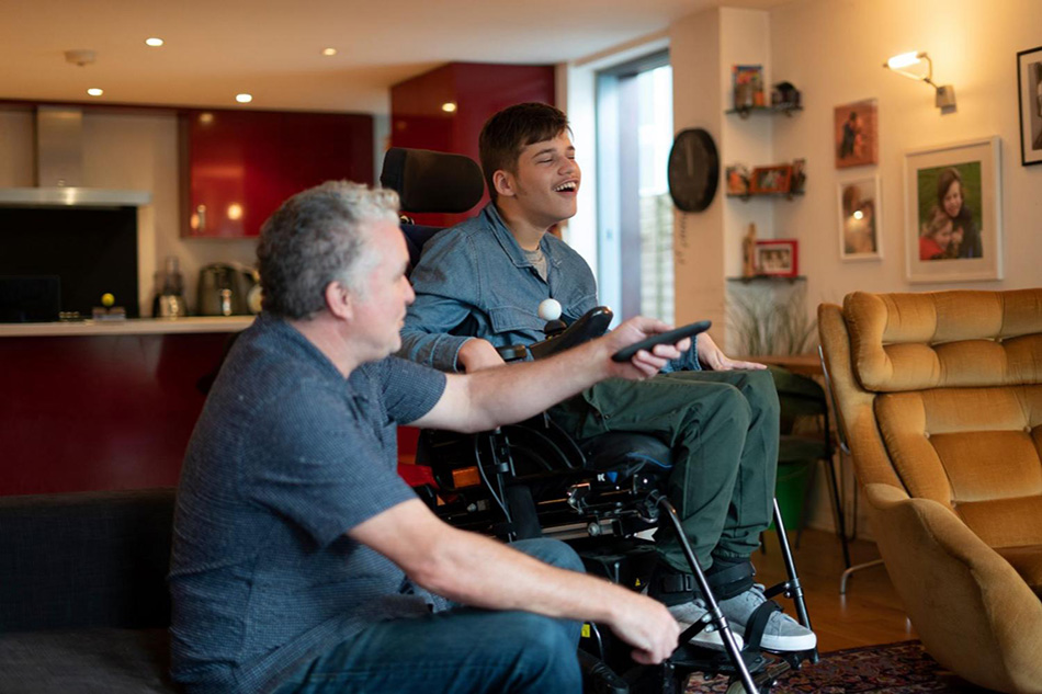 Young man seated in wheelchair in home beside support worker