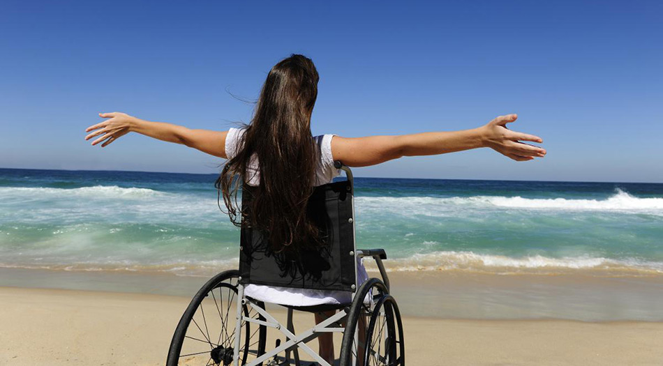 Womain sitting in a wheelchair at the beach with her arms open wide as she looks at the sea