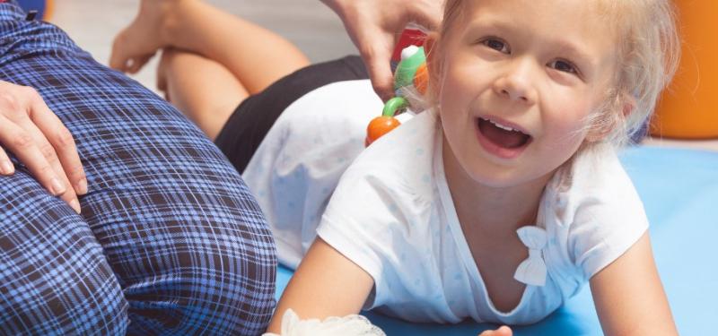 smiling preschool aged girl lays on her belly next to a caretaker and toys