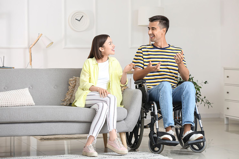 A young man is sitting in a wheelchair next to a young woman on a couch