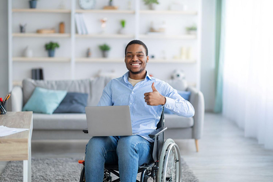 Male seated in wheelchair with laptop showing thumb up gesture, smiling at camera