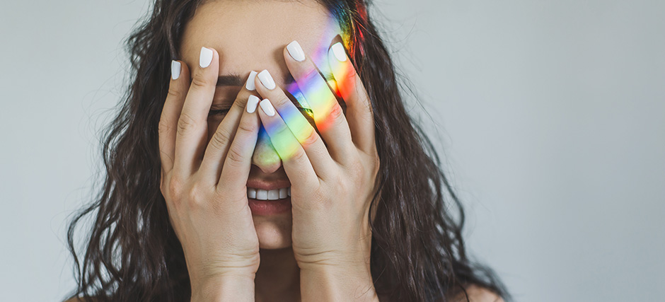 A young woman covering her face with her hands and a rainbow of light is shinging aross her hands