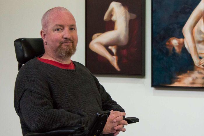 Andrew Grant sits in front of his paintings and is smiling