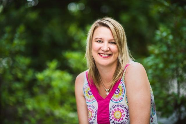 Professional photo of Jessica. She is sitting outside with trees behind her, and she smiles and is wearing a pink floral print shirt