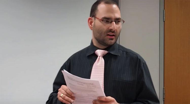 Paul standing speaking holding his notes and wearing a pink tie