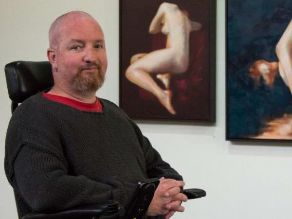 Andrew Grant sits in front of his paintings and is smiling