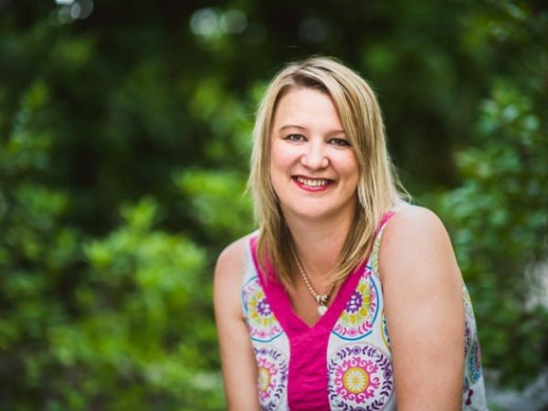 Professional photo of Jessica. She is sitting outside with trees behind her, and she smiles and is wearing a pink floral print shirt